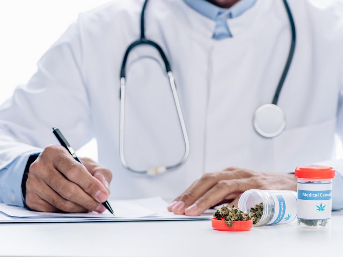 Stock photo of doctor sitting at desk and writing with medicine containers containing dried cannabis on the table