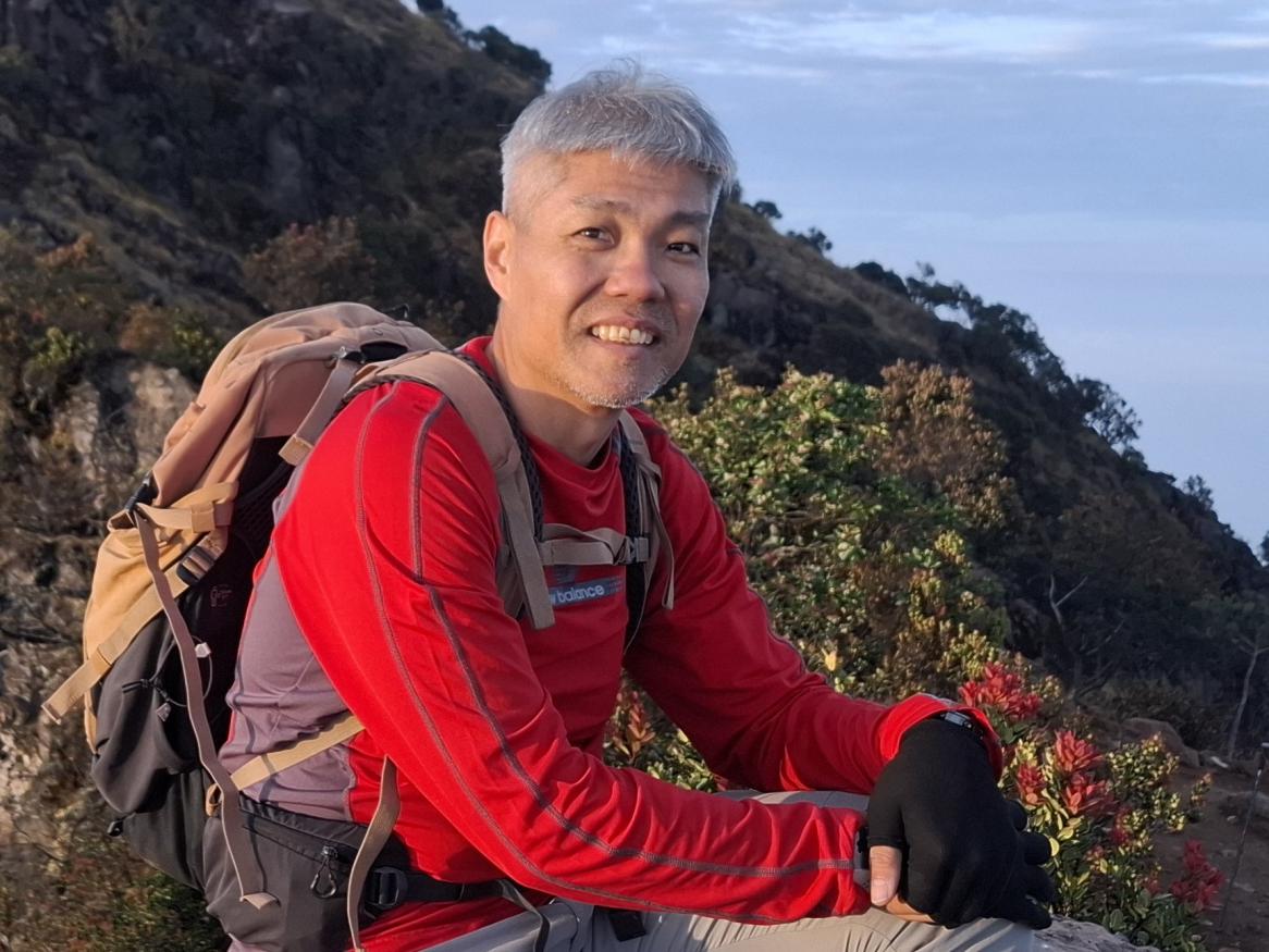 A portrait of Sam Ng sitting on a rock, on a mountain